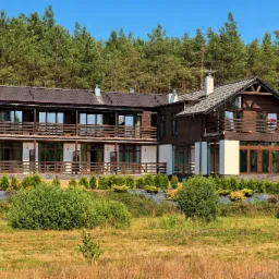 Dwupiętrowy budynek z ciemnego drewna z balkonami i jasnymi elementami elewacji, otoczony zielenią i drzewami iglastymi.