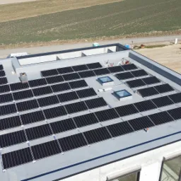 Widok z góry na instalację paneli słonecznych na płaskim dachu budynku w trakcie montażu, z widocznym żurawiem budowlanym w tle.