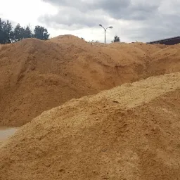 Góra jasnych trocin i zrębków drzewnych na betonowym placu, widoczne drzewa w tle, pochmurne niebo