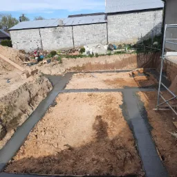 Wykopane fundamenty z zalanym betonem, widok z g&oacute;ry na plac budowy z ziemią i szarym betonem, w tle budynek gospodarczy z szarej cegły.