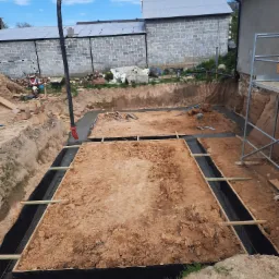 Wykop pod fundamenty domu jednorodzinnego z deskowaniem i hydroizolacją, częściowo zalany betonem z pompy, widoczny skuter w tle.