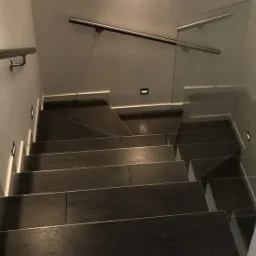 Schody wyłożone ciemnymi płytkami z podświetleniem LED w ścianie, balustrada ze stali nierdzewnej z elementami szklanymi, widok z góry.