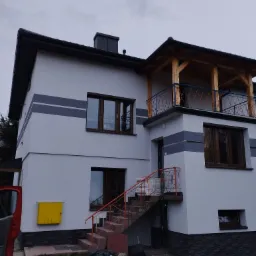 Dwupiętrowy dom z jasnoszarą elewacją, ciemnymi pasami dekoracyjnymi, drewnianym balkonem z niebieską balustradą i schodami z pomarańczową barierką, widok z zewnątrz na pochmurny dzień.