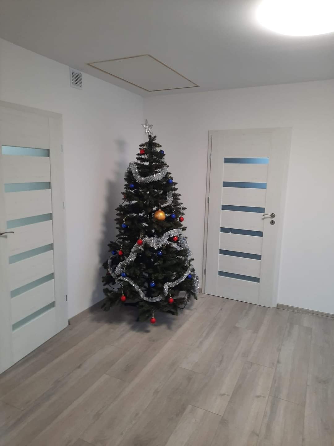 Wnętrze pokoju z szarą podłogą imitującą drewno, białe ściany i białe drzwi z poziomymi wgłębieniami, w rogu stoi choinka z srebrnym łańcuchem i kolorowymi bombkami, sufit z klapą rewizyjną.
