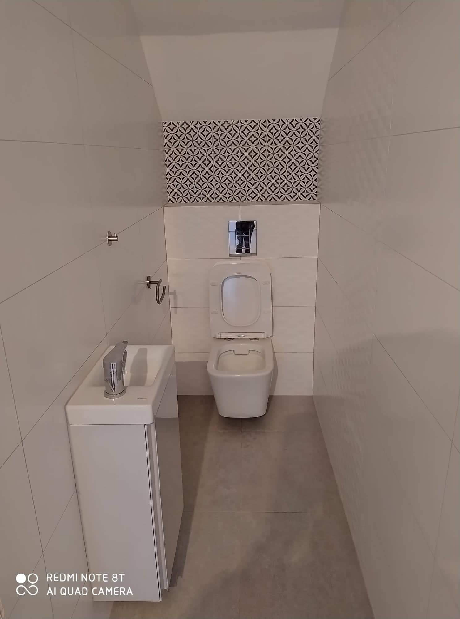 Mała, nowoczesna toaleta z umywalką z szafką, białe płytki ścienne z szarymi fugami ułożone poziomo, dekoracyjny pas płytek nad spłuczką, szara podłoga, otwarte siedzisko toalety.