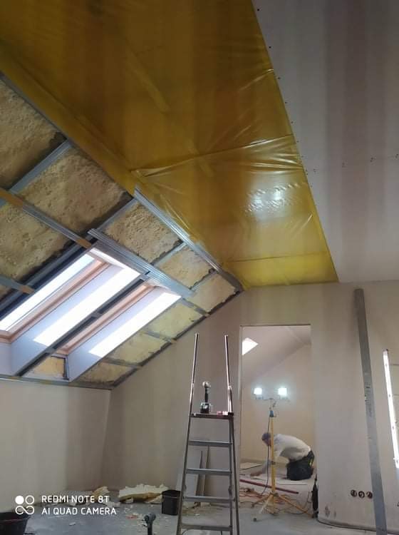 Poddasze w trakcie ocieplania z widoczną konstrukcją metalową, izolacją z wełny mineralnej, folią paroizolacyjną w kolorze żółtym, oknami dachowymi oraz pracownikiem na drabinie przygotowującym się...