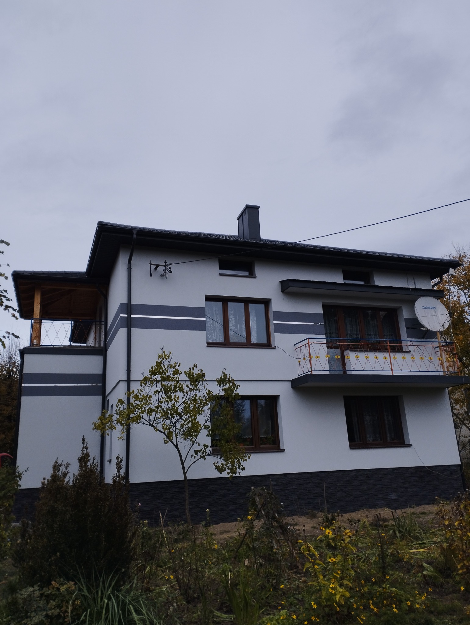 Dwukondygnacyjny dom z szarą elewacją, ciemnymi pasami i balkonem z ozdobną balustradą, widoczny z poziomu ogrodu w pochmurny dzień.