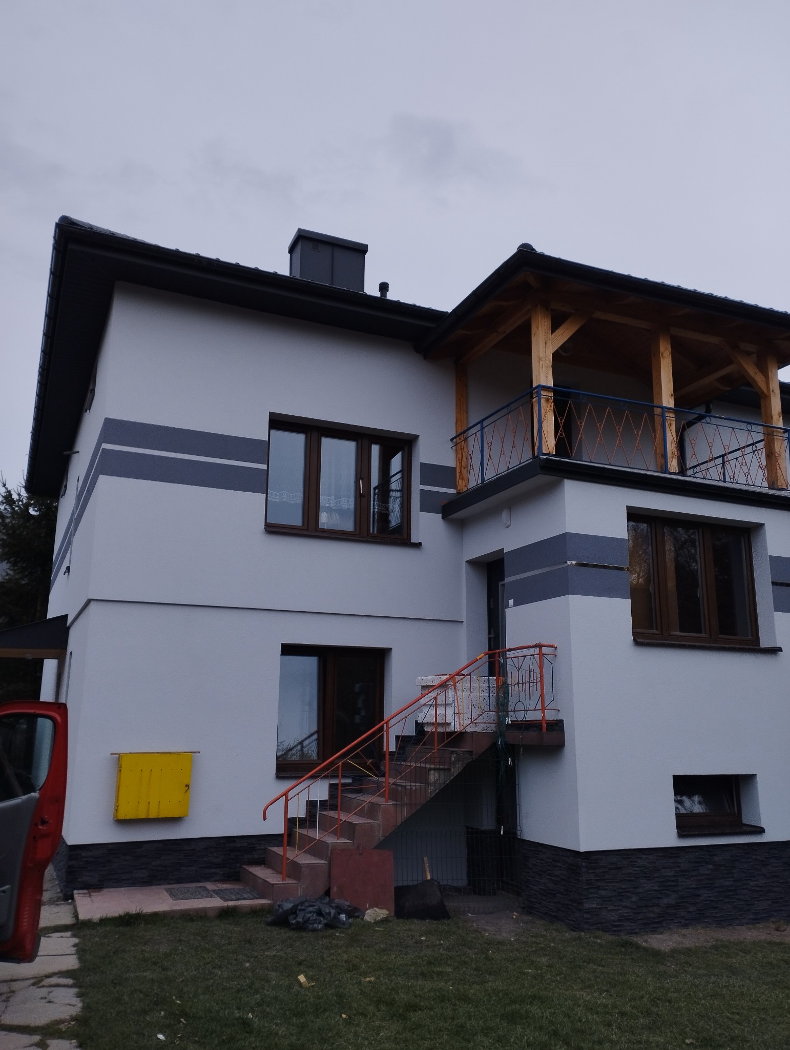 Dwupiętrowy dom z jasnoszarą elewacją, ciemnymi pasami dekoracyjnymi, drewnianym balkonem z niebieską balustradą i schodami z pomarańczową barierką, widok z zewnątrz na pochmurny dzień.