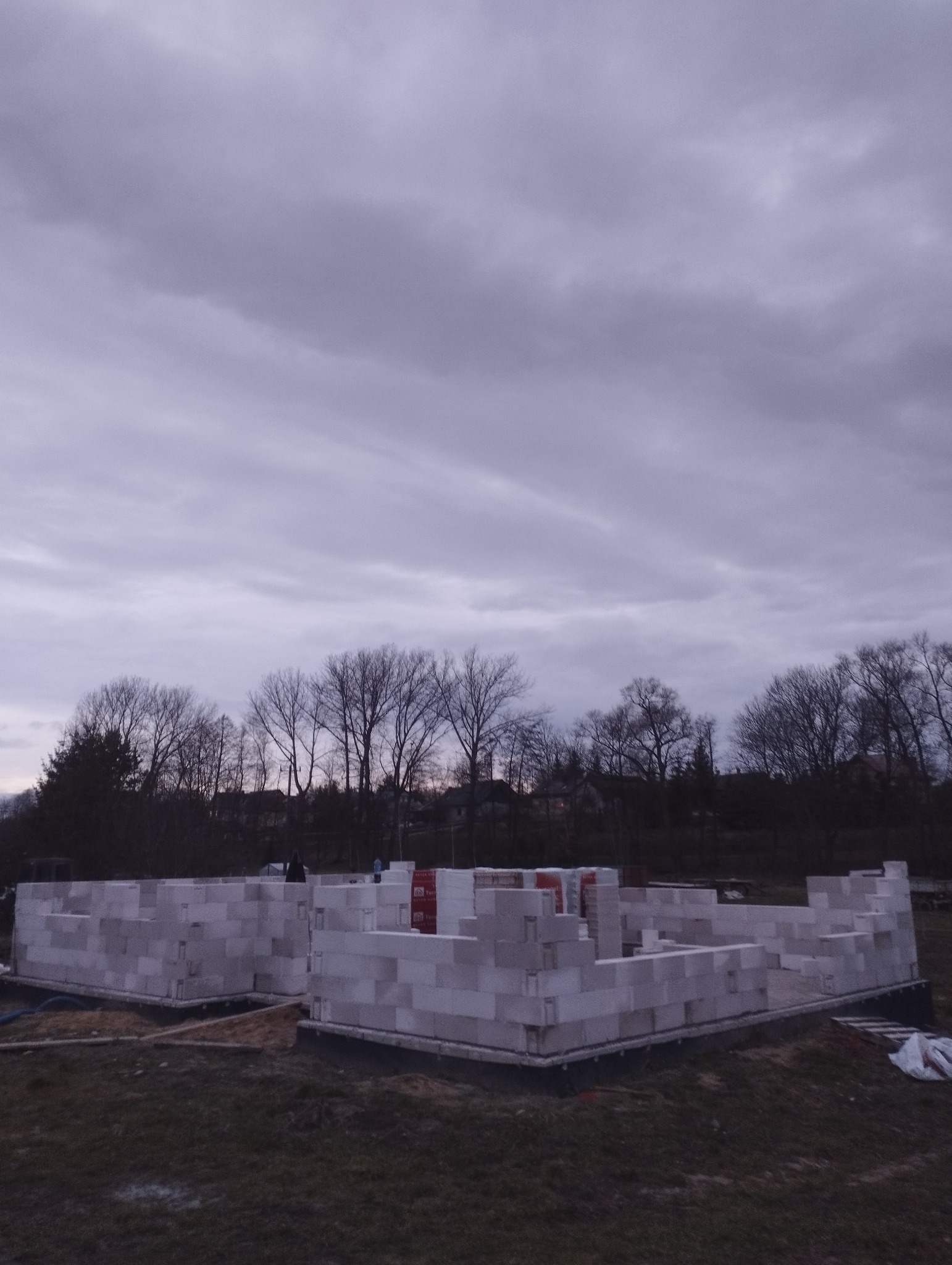 Fundamenty domu z betonu komórkowego w trakcie budowy, widok na szkielet ścian parteru na tle pochmurnego nieba i drzew bez liści.