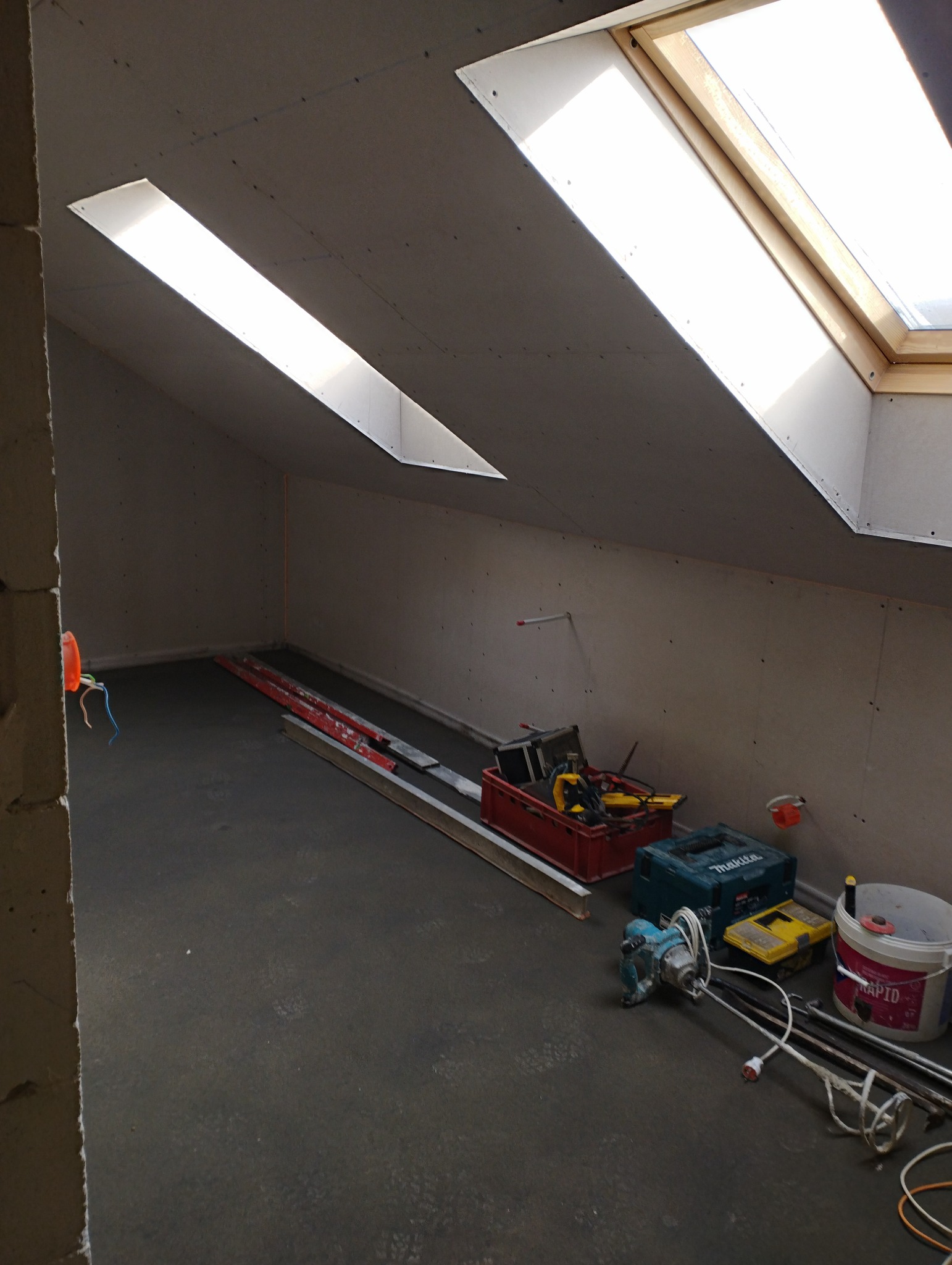 Poddasze w trakcie wykańczania, widoczne okna dachowe, ściany pokryte płytami gipsowo-kartonowymi, narzędzia i materiały budowlane na podłodze, w tym wiertarka i mieszadło do zaprawy.