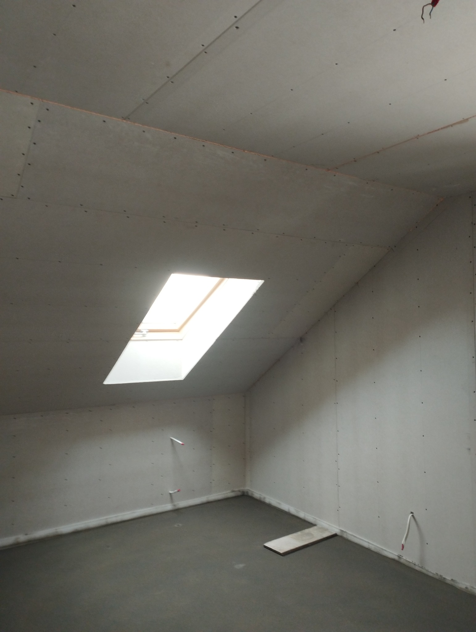 Poddasze w trakcie wykańczania: zamontowane płyty gipsowo-kartonowe na ścianach i suficie, widoczne łączenia płyt, okno dachowe w skosie sufitu, wylewka na podłodze, wystające kable elektryczne.