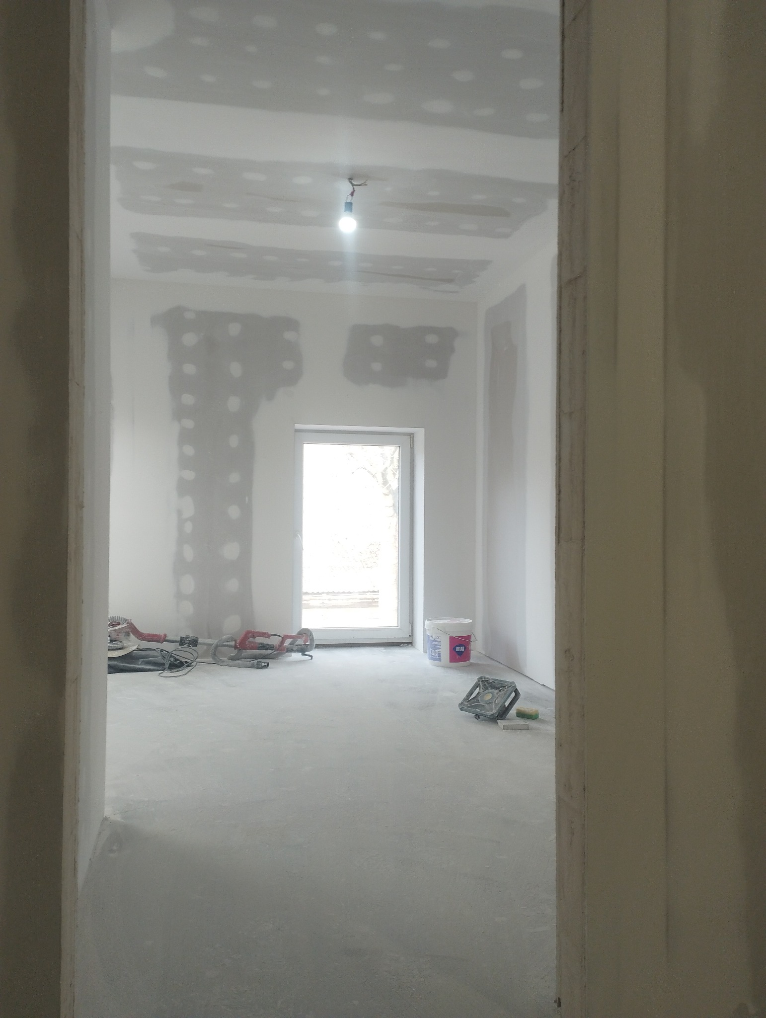 Wnętrze w trakcie prac przygotowawczych do wykończenia w Miechowie: ściany z płyt gipsowo-kartonowych z widocznymi łączeniami szpachlowanymi, narzędzia szlifierskie na podłodze, wiadro farby przy...