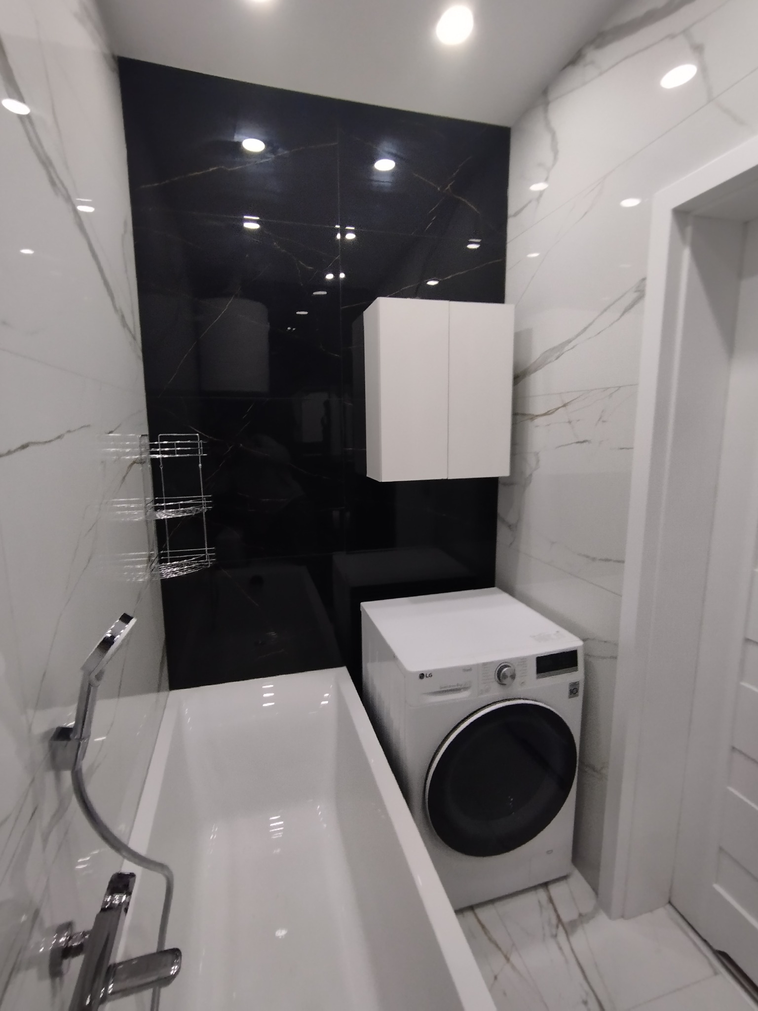 Nowoczesna łazienka z wanną, czarnymi płytkami imitującymi marmur na ścianie, białą szafką wiszącą i pralką LG.