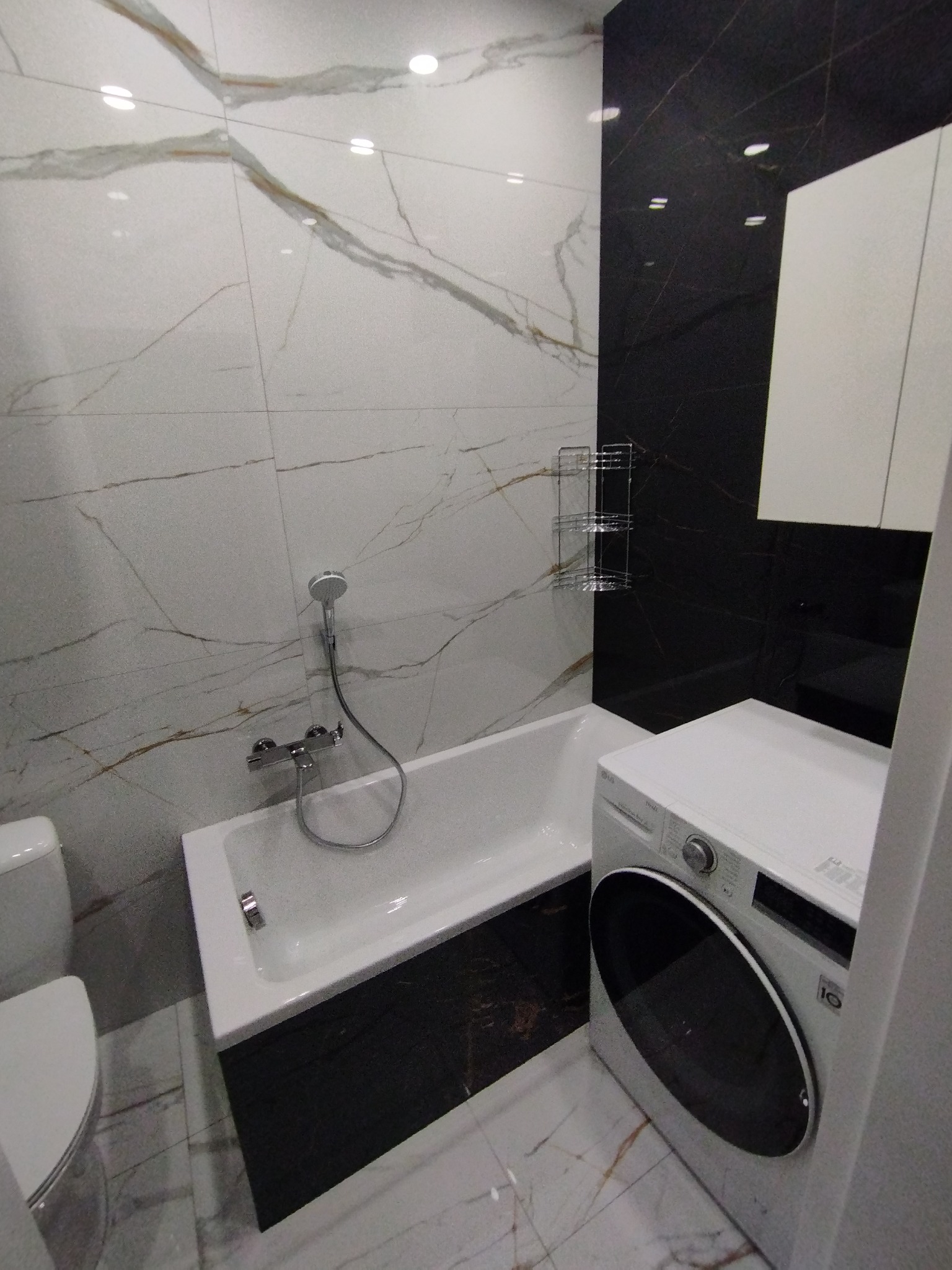 Wnętrze łazienki z wanną obudowaną czarnymi płytkami, białą umywalką i pralką LG, wykończone białymi płytkami z marmurkowym wzorem na ścianach.