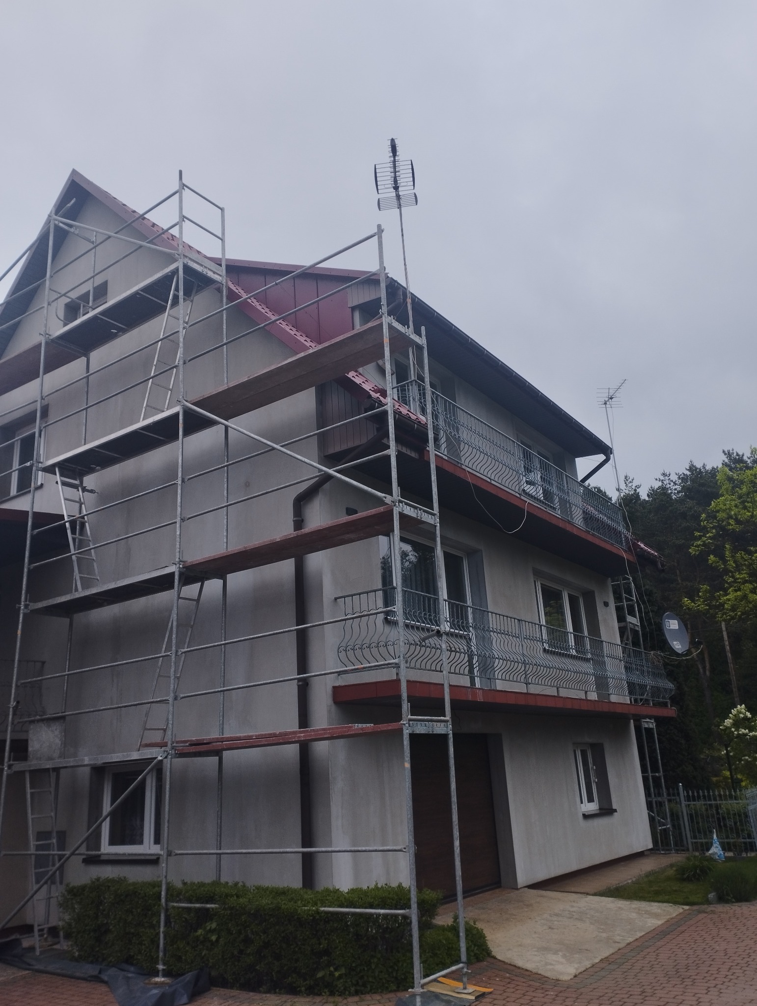 Dwupiętrowy dom z balkonami w trakcie prac elewacyjnych, otoczony rusztowaniem, widoczna antena na dachu.