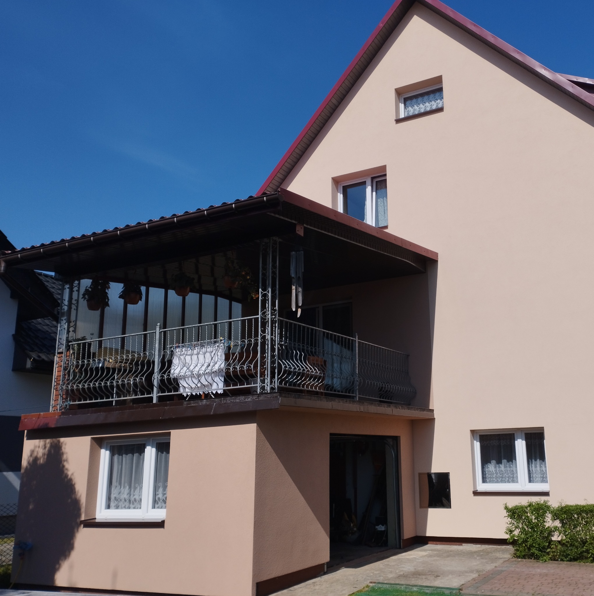 Dwupiętrowy dom z beżową elewacją, balkonem z metalową balustradą i zadaszeniem, widok z dołu w słoneczny dzień.