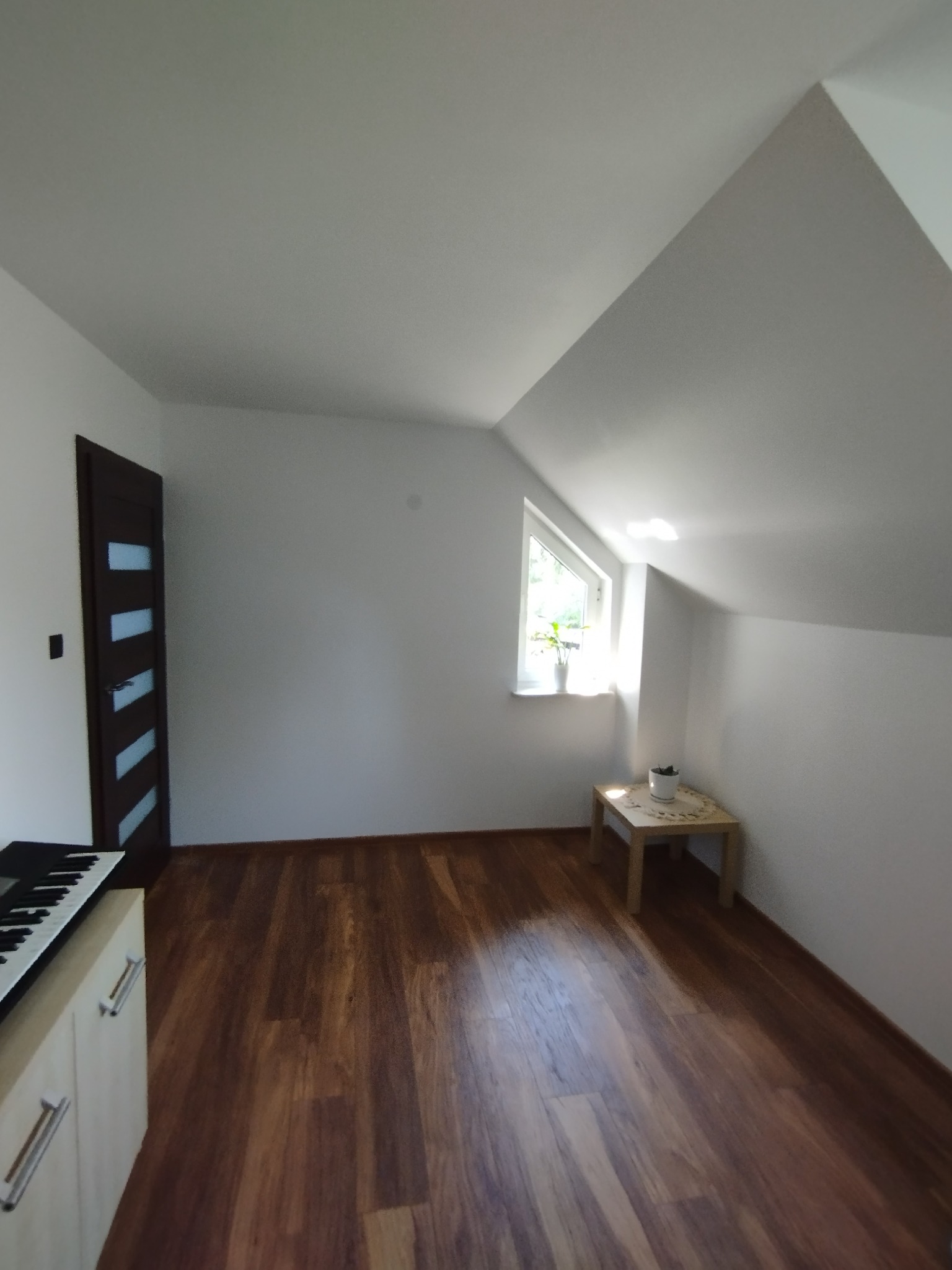 Pokój na poddaszu z drewnianą podłogą, oknem z rośliną i stolikiem, częściowo widoczny instrument klawiszowy po lewej stronie.