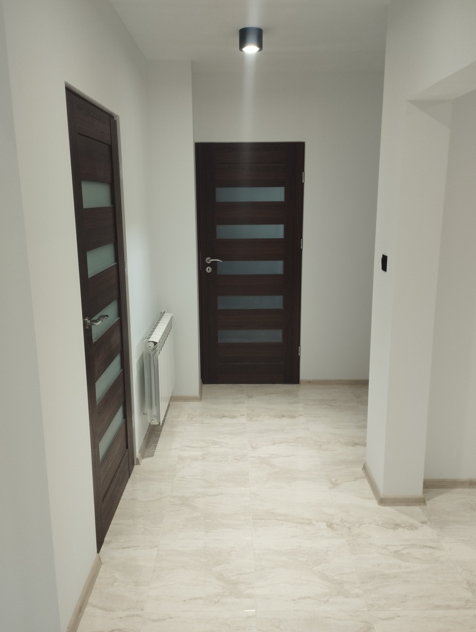 Korytarz po remoncie z dwiema ciemnymi, fornirowanymi drzwiami z przeszkleniami, jasną marmurową podłogą i białymi ścianami oraz grzejnikiem.