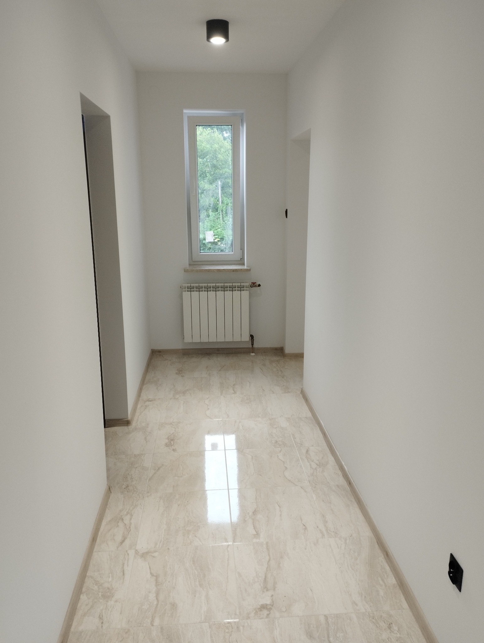 Wykończony korytarz w jasnych barwach z marmurową podłogą, białymi ścianami, grzejnikiem pod oknem i punktowym oświetleniem sufitowym.