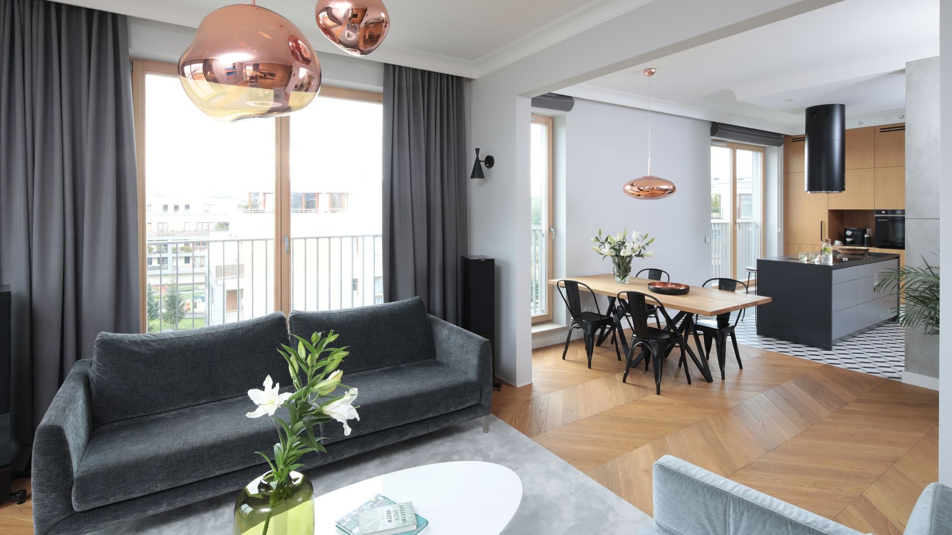 Jasne, przestronne wnętrze mieszkania z parkietem w jodełkę, szarą sofą i miedzianymi lampami wiszącymi, widok na balkon i kuchnię z wyspą.