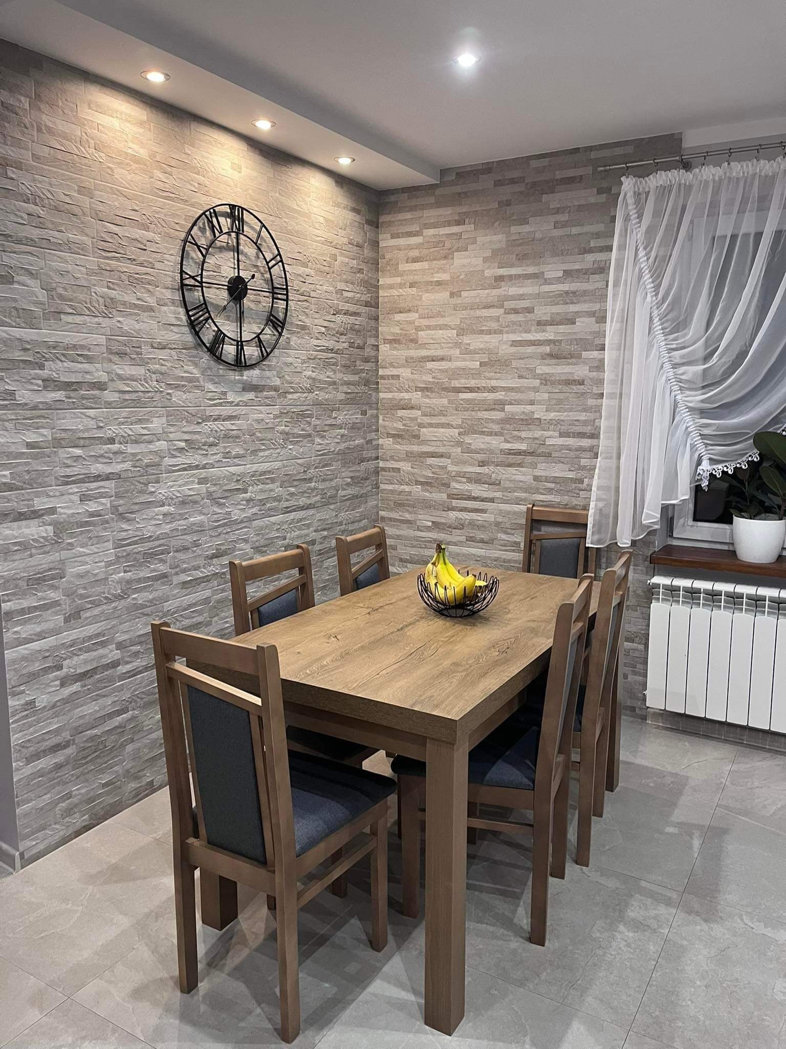 Jadalnia z drewnianym stołem i krzesłami na tle ściany wyłożonej płytkami imitującymi kamień, z dekoracyjnym zegarem na ścianie.