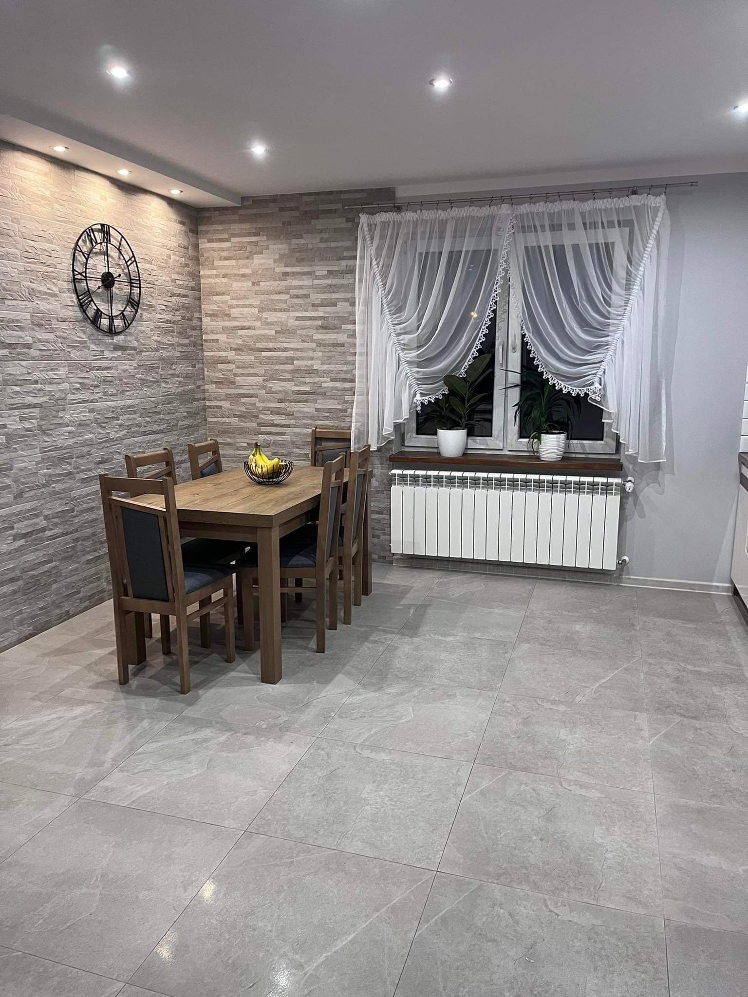Jasne wnętrze jadalni z drewnianym stołem i krzesłami, szarą kamienną ścianą i podłogą, ozdobnym zegarem oraz oknem z białymi firankami i roślinami doniczkowymi.