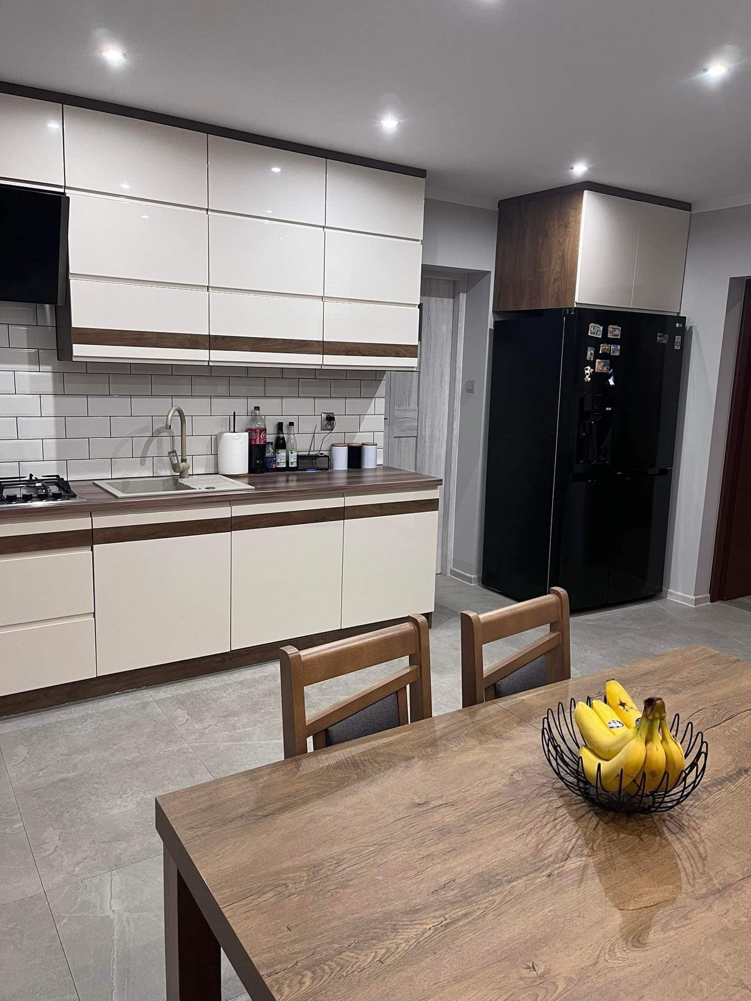 Nowoczesna kuchnia z białymi szafkami, drewnianymi akcentami, czarną lodówką side-by-side i stołem z owocami w metalowej misie.