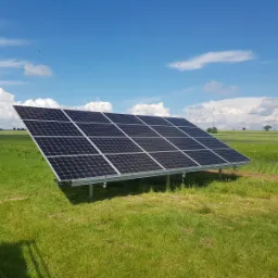 Panel słoneczny na metalowej konstrukcji, ustawiony na polu z zieloną trawą, w tle błękitne niebo z białymi chmurami i turbiny wiatrowe.