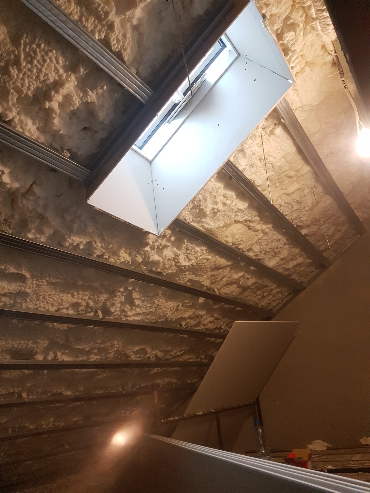 Poddasze z oknem dachowym w trakcie ocieplania pianą PUR, widoczne metalowe profile konstrukcyjne i płyta gipsowo-kartonowa oparta o ścianę.