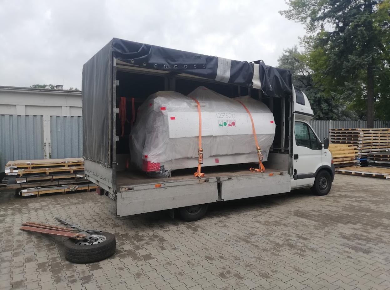 Biały bus z otwartą plandeką przewozi zabezpieczoną folią maszynę, przymocowaną pomarańczowymi pasami transportowymi. Obok leżą opony i narzędzia, a w tle widać palety.