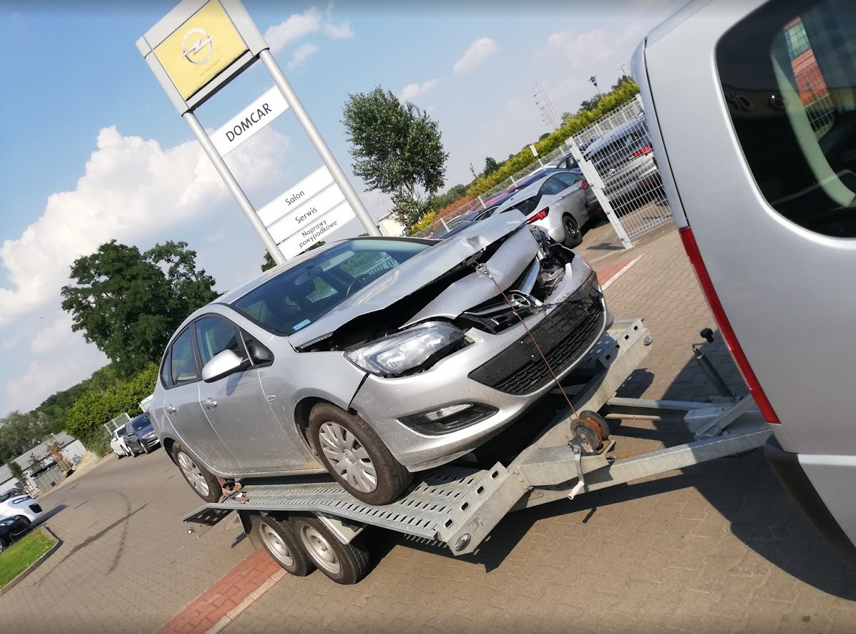 Uszkodzony srebrny Opel Astra na lawecie, widoczny budynek salonu samochodowego Domcar w tle.