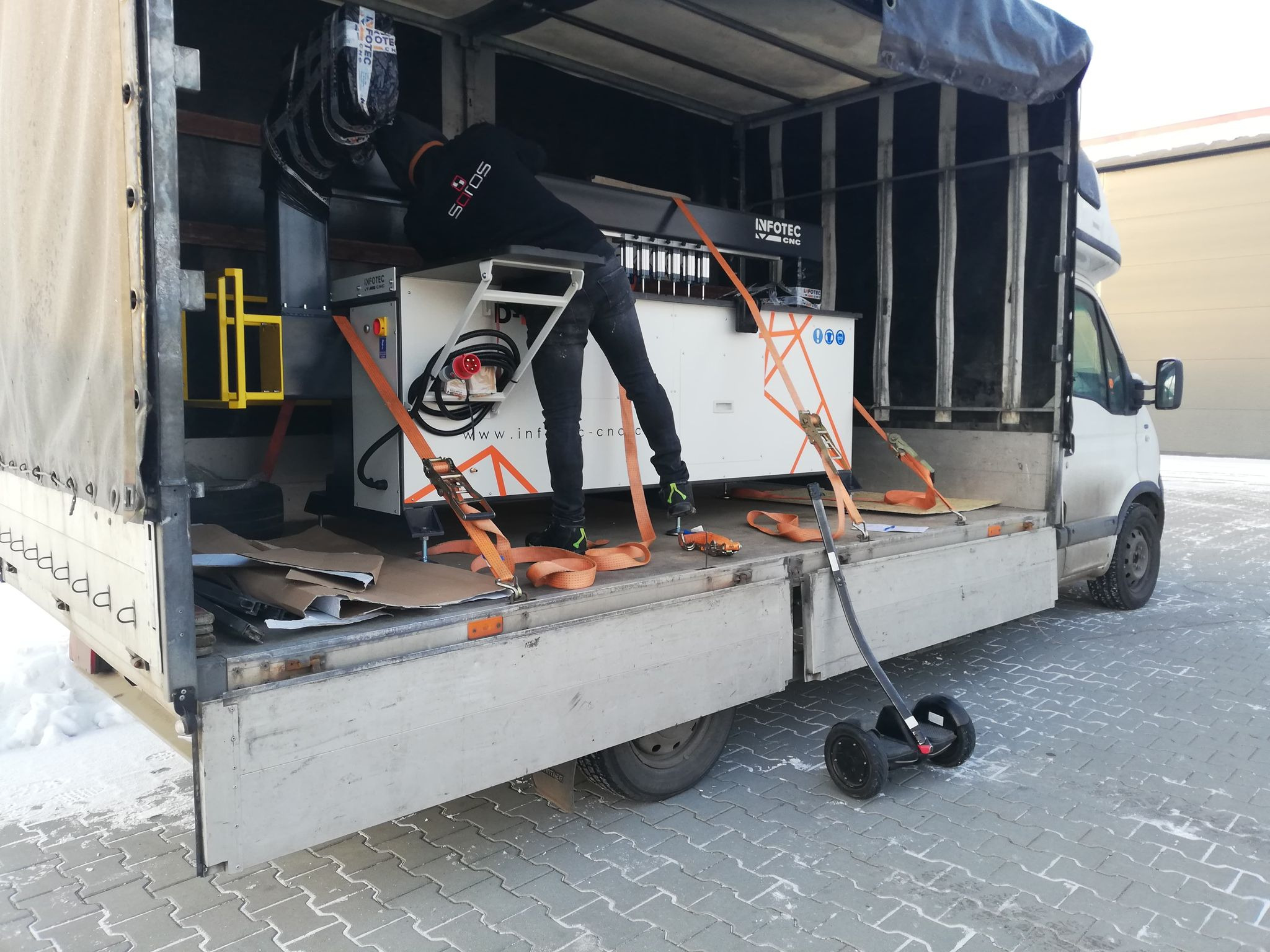 Załadunek maszyny CNC Infotec do busa transportowego, zabezpieczonej pomarańczowymi pasami, widoczny pracownik w czarnej bluzie Sojos i elektryczny wózek transportowy na zewnątrz.