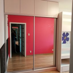 Mebel Pro Dariusz Pieron - Biała szafa z przesuwnymi lustrzanymi drzwiami, z nadstawką, odbijająca wnętrze pokoju z różową ścianą i drewnianą podłogą. W lustrze widoczna osoba robiąca zdjęcie.