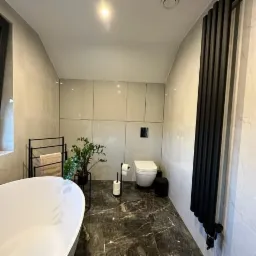 Nowoczesna łazienka z marmurową podłogą, wolnostojąca wanna, grzejnik żeberkowy w kolorze czarnym, jasne płytki ścienne i roślina doniczkowa na czarnym stojaku.