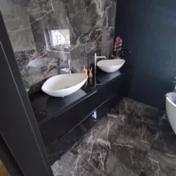 Nowoczesna łazienka z dwoma białymi umywalkami nablatowymi na czarnym blacie z marmuru, ściany wyłożone marmurem w odcieniach szarości i czerni, w tle widoczna ściana z pionowymi panelami i kwiat...