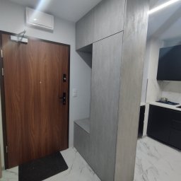 Mebel Pro Dariusz Pieron - Szara zabudowa meblowa w przedpokoju z siedziskiem i szafkami, dopasowana do wysokości pomieszczenia, z drewnianymi drzwiami wejściowymi i widoczną częścią kuchni w tle.