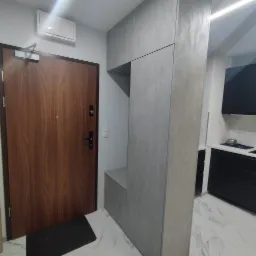 Szara zabudowa meblowa w przedpokoju z siedziskiem i szafkami, dopasowana do wysokości pomieszczenia, z drewnianymi drzwiami wejściowymi i widoczną częścią kuchni w tle.