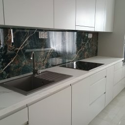 Mebel Pro Dariusz Pieron - Nowoczesna kuchnia z białymi szafkami, blatem imitującym marmur i panelem szklanym nad blatem z motywem ciemnego marmuru z jasnymi żyłami. Widoczny zlew w kolorze grafitowym i płyta indukcyjna.