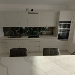 Mebel Pro Dariusz Pieron - Nowoczesna kuchnia na wymiar z białymi frontami, blatem imitującym marmur i zielonym panelem nad blatem, widok zza stołu z marmurowym blatem.