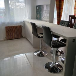 Mebel Pro Dariusz Pieron - Wysoki, biały blat z marmurowym wzorem, wsparty na szafkach, z trzema nowoczesnymi, czarnymi hokerami na chromowanych nogach. W tle okno z firanką i grzejnik żebrowy.