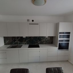 Mebel Pro Dariusz Pieron - Jasna, nowoczesna zabudowa kuchenna z frontami bez uchwytów, blatem w kolorze marmuru i ciemnozielonym panelem nad blatem, z wbudowanym piekarnikiem i płytą grzewczą.