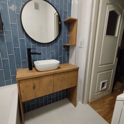 Mebel Pro Dariusz Pieron - Nowoczesna łazienka z drewnianą szafką pod umywalką nablatową, czarną armaturą i okrągłym lustrem na ścianie z niebieskich płytek imitujących cegłę.