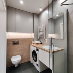 Mebel Pro Dariusz Pieron - Nowoczesna łazienka z zabudową nad toaletą, pralką pod blatem drewnianym i szklanym panelem prysznicowym, szare płytki i oświetlenie LED.