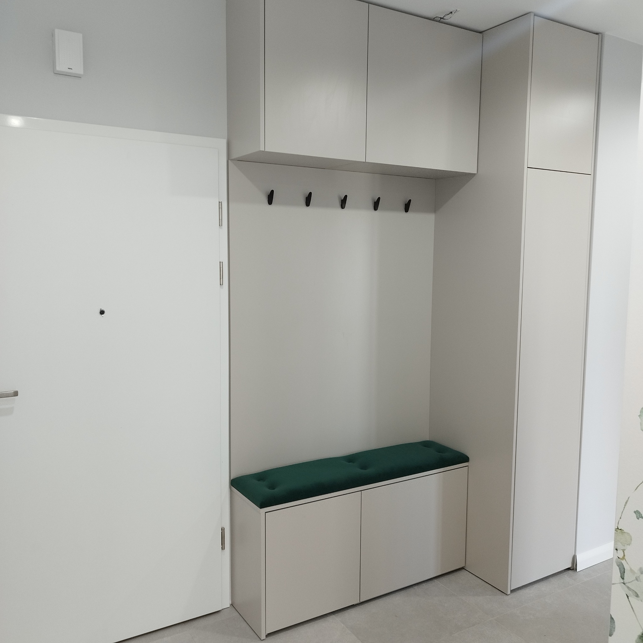 Jasnoszara zabudowa przedpokoju z szafkami górnymi, wieszakami i siedziskiem z zieloną tapicerką, widok na białe drzwi wejściowe.