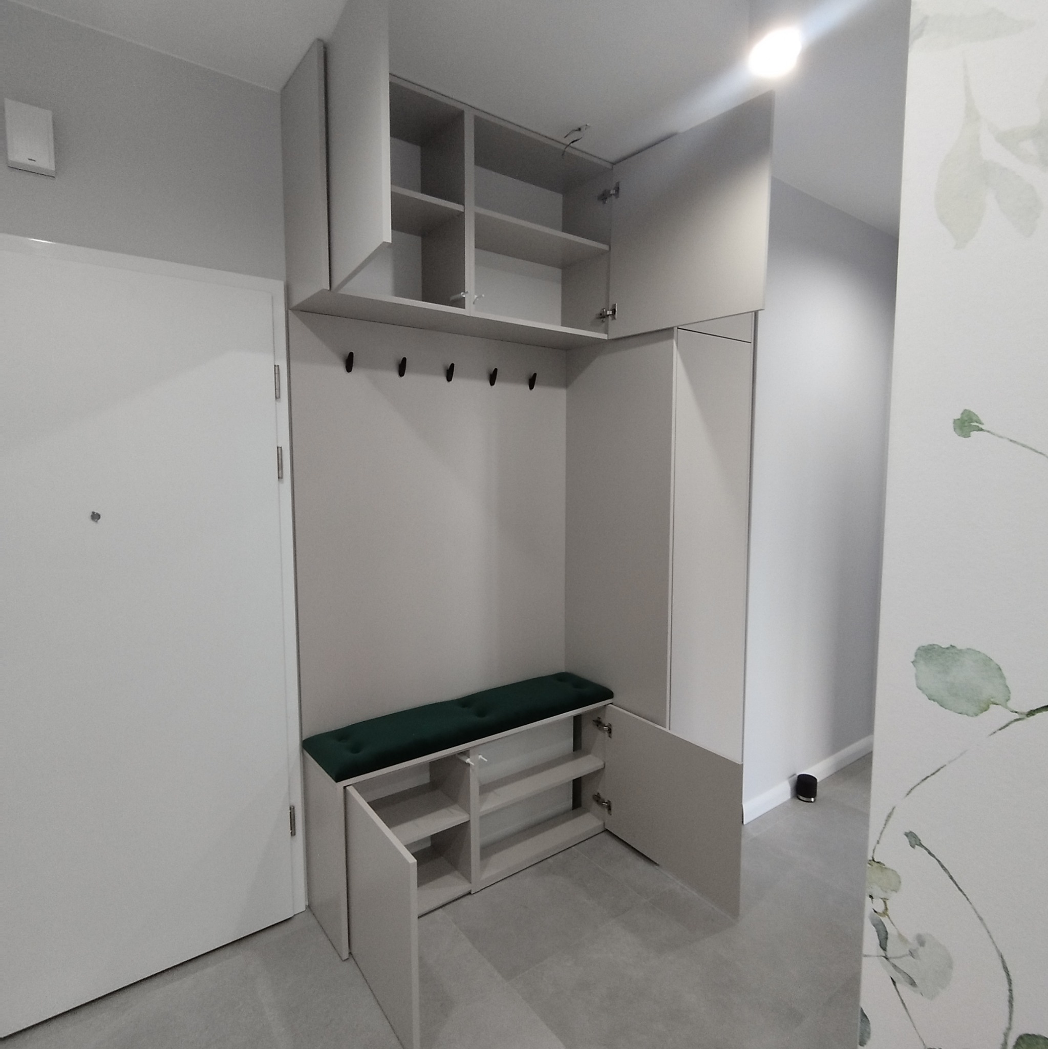 Jasnoszara zabudowa przedpokoju z siedziskiem z zieloną tapicerką, otwartymi szafkami na buty i wieszakami na ubrania, uzupełniona górnymi szafkami z półkami.