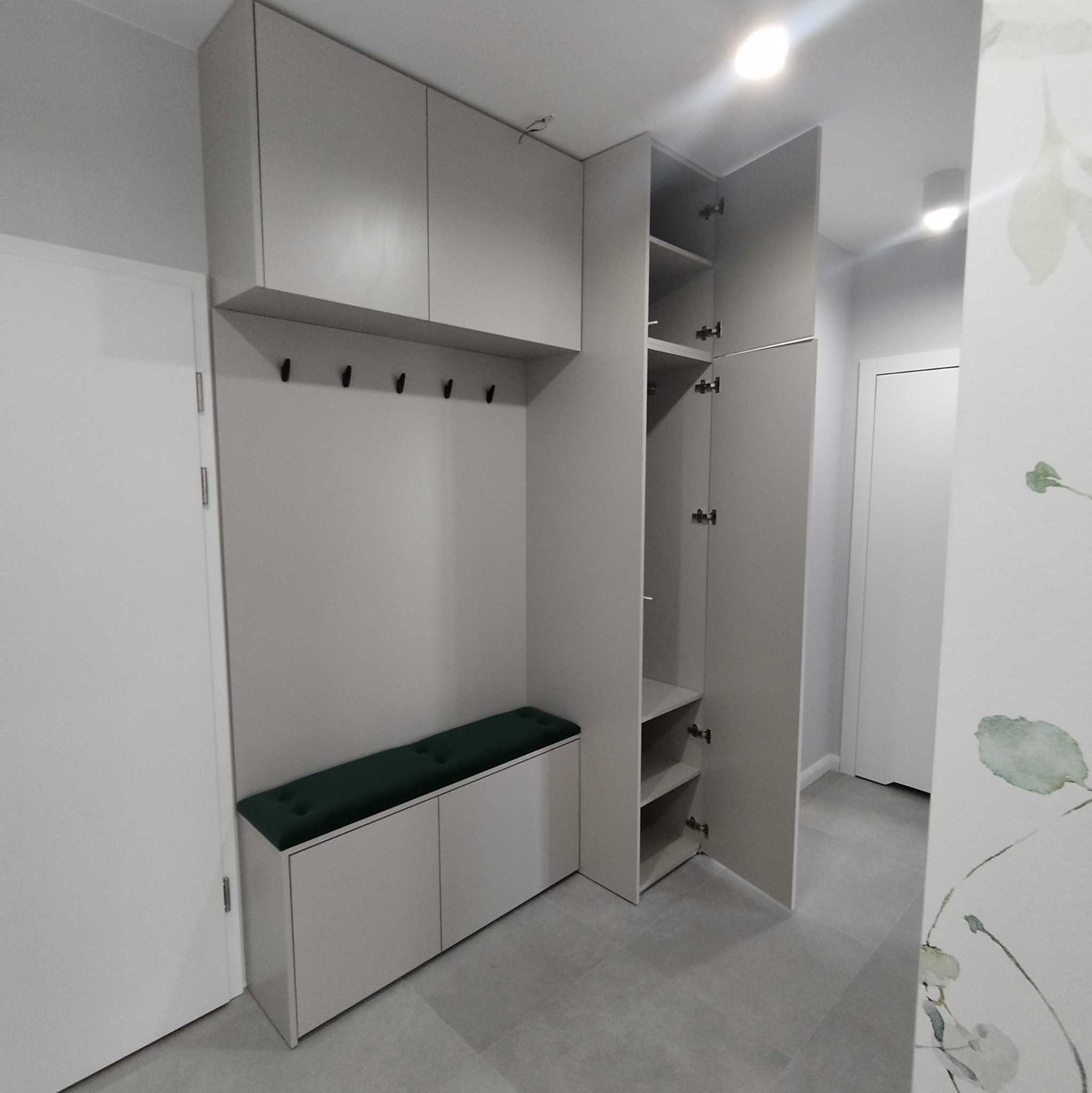 Jasnoszara zabudowa przedpokoju z szafkami górnymi, wieszakami, siedziskiem z zieloną poduszką i otwartą szafą z półkami, widok na szarą podłogę i fragment ściany z motywem roślinnym.
