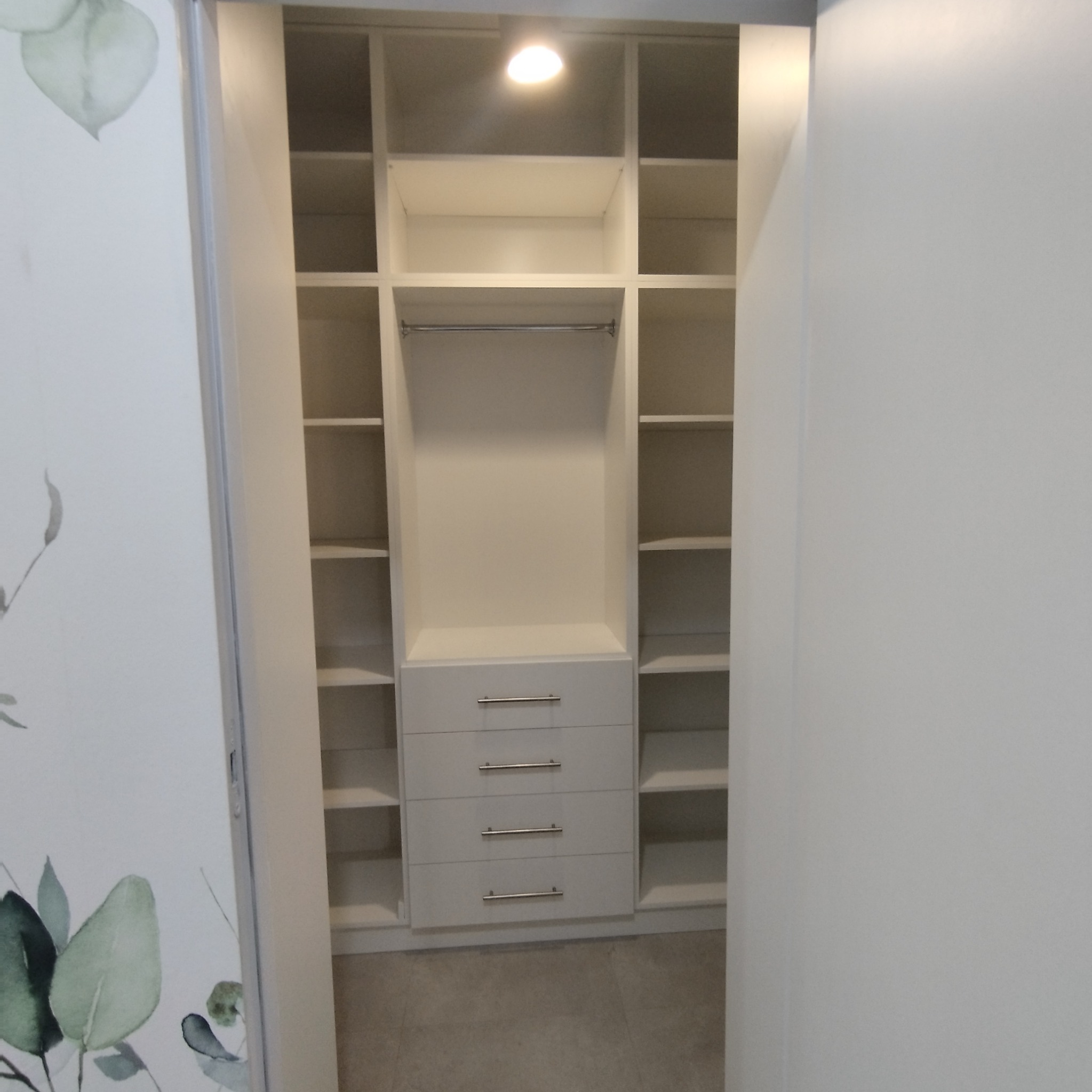 Wnętrze białej szafy z półkami i czterema szufladami, oświetlone punktowym światłem, widoczne przez częściowo otwarte drzwi z delikatnym motywem roślinnym.