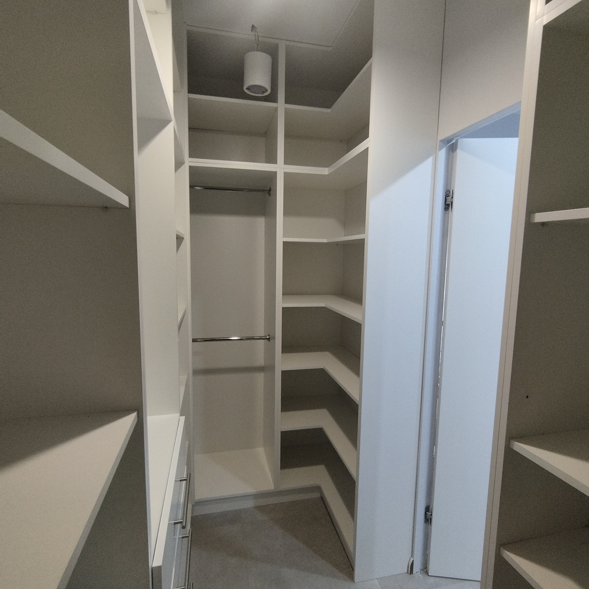 Wnętrze garderoby z białymi półkami, drążkami na ubrania i szufladami, oświetlenie sufitowe w neutralnej kolorystyce.