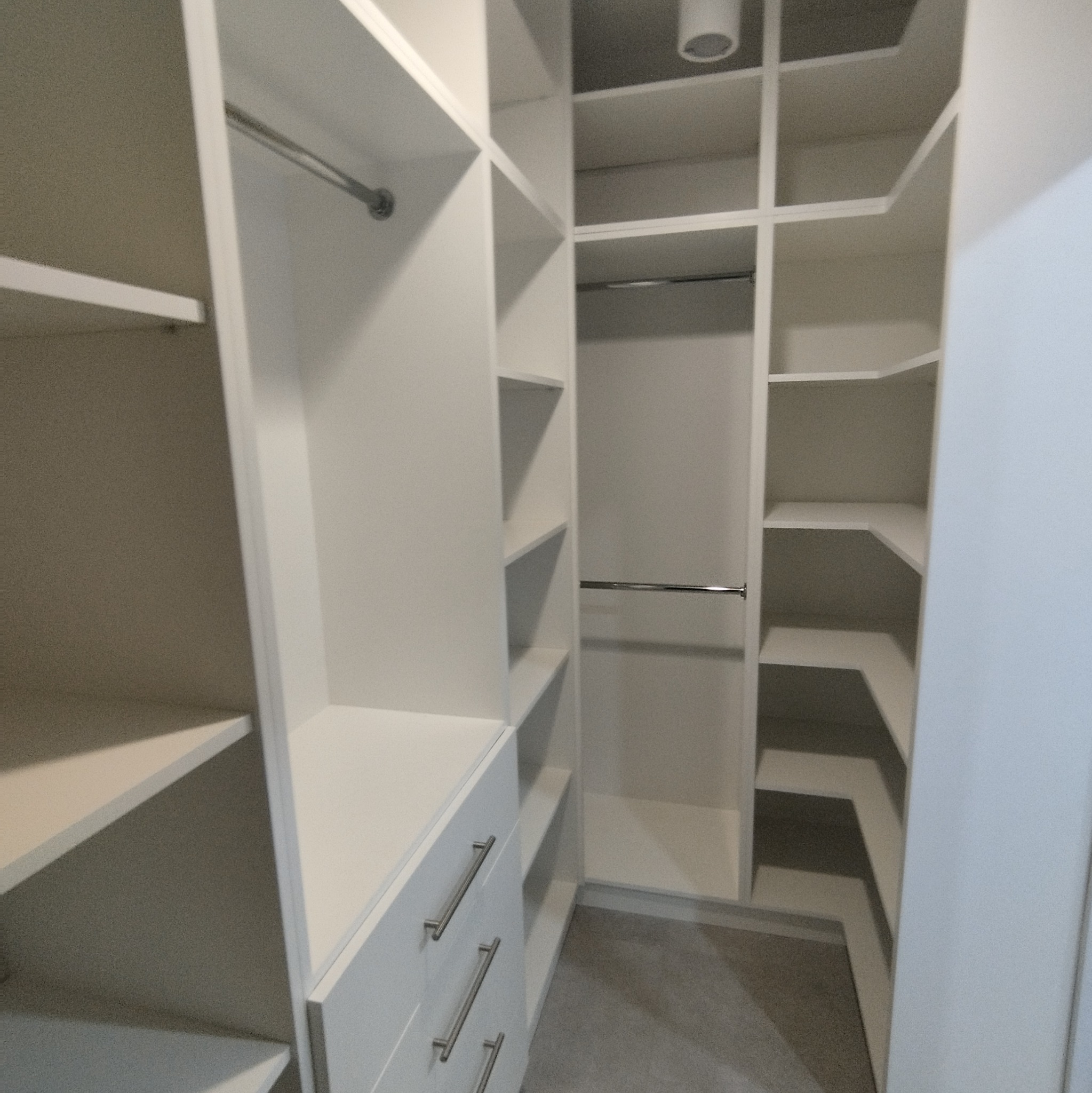 Wnętrze białej, nowoczesnej garderoby z półkami, szufladami z podłużnymi uchwytami i drążkami na ubrania, oświetlenie sufitowe.