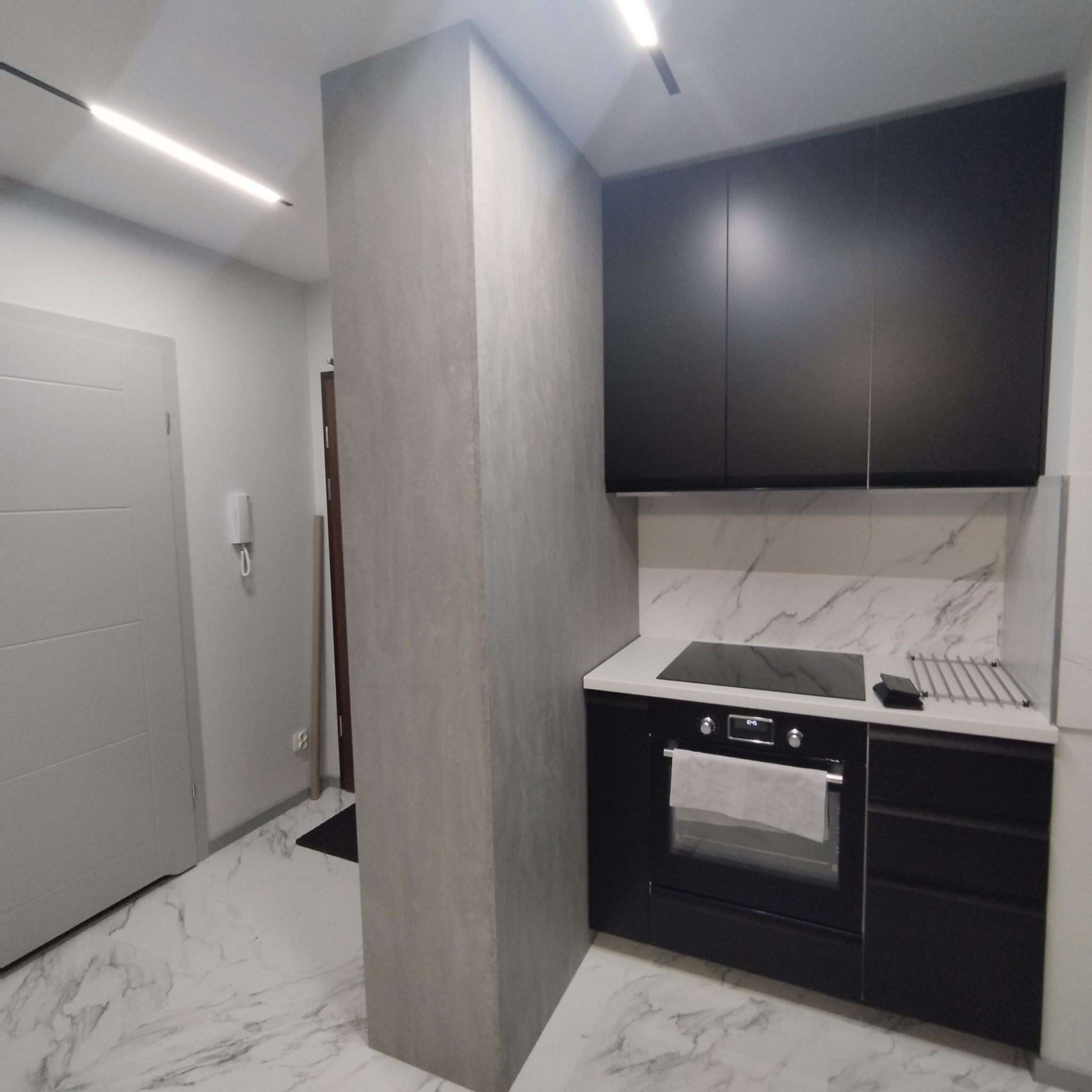 Nowoczesna kuchnia z czarnymi szafkami i białym blatem, marmurowa podłoga i ściana nad blatem, widoczny fragment kolumny w kolorze betonu.